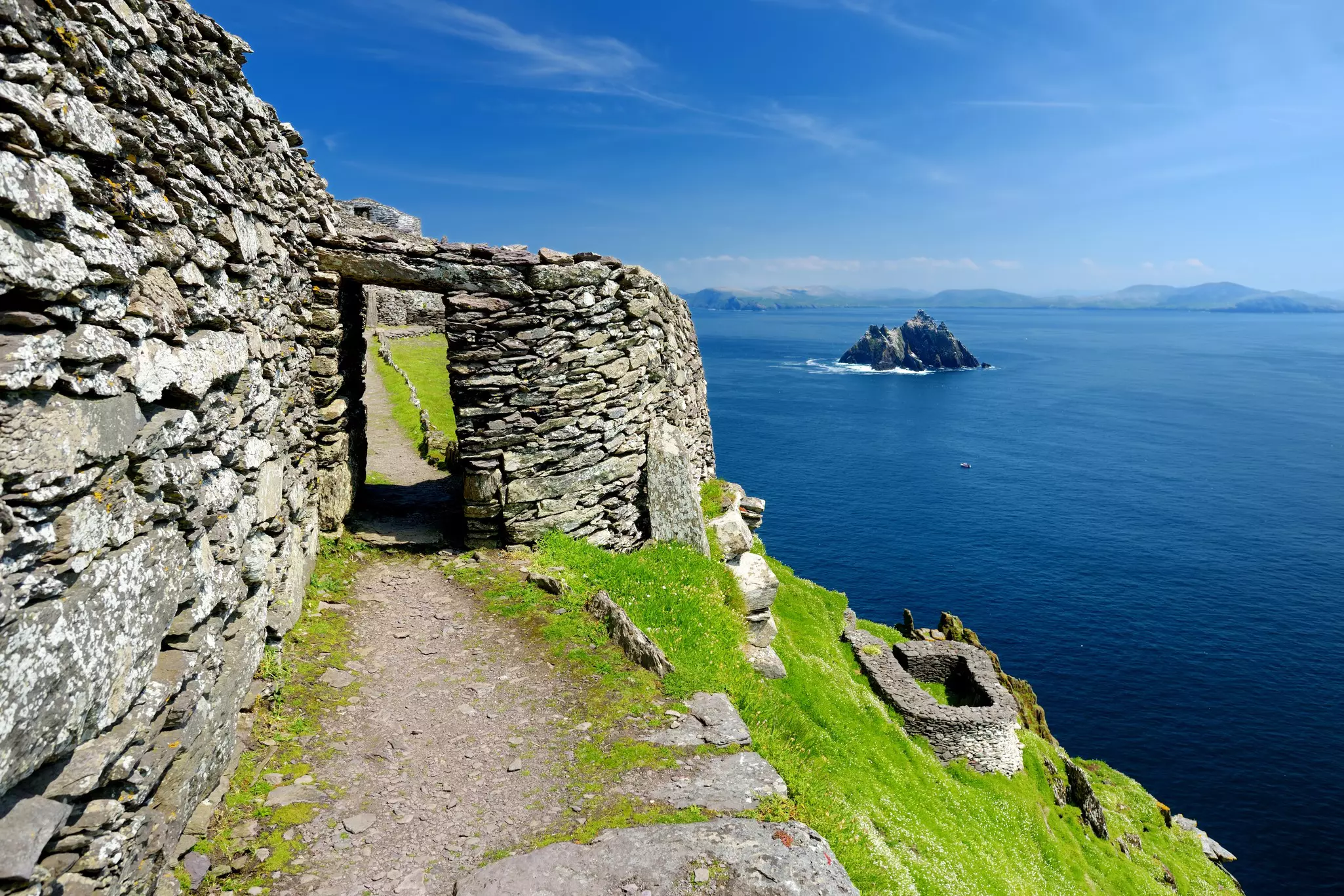 A rocky stone wall at the edge of an island with a rocky islet out at sea in the distance.