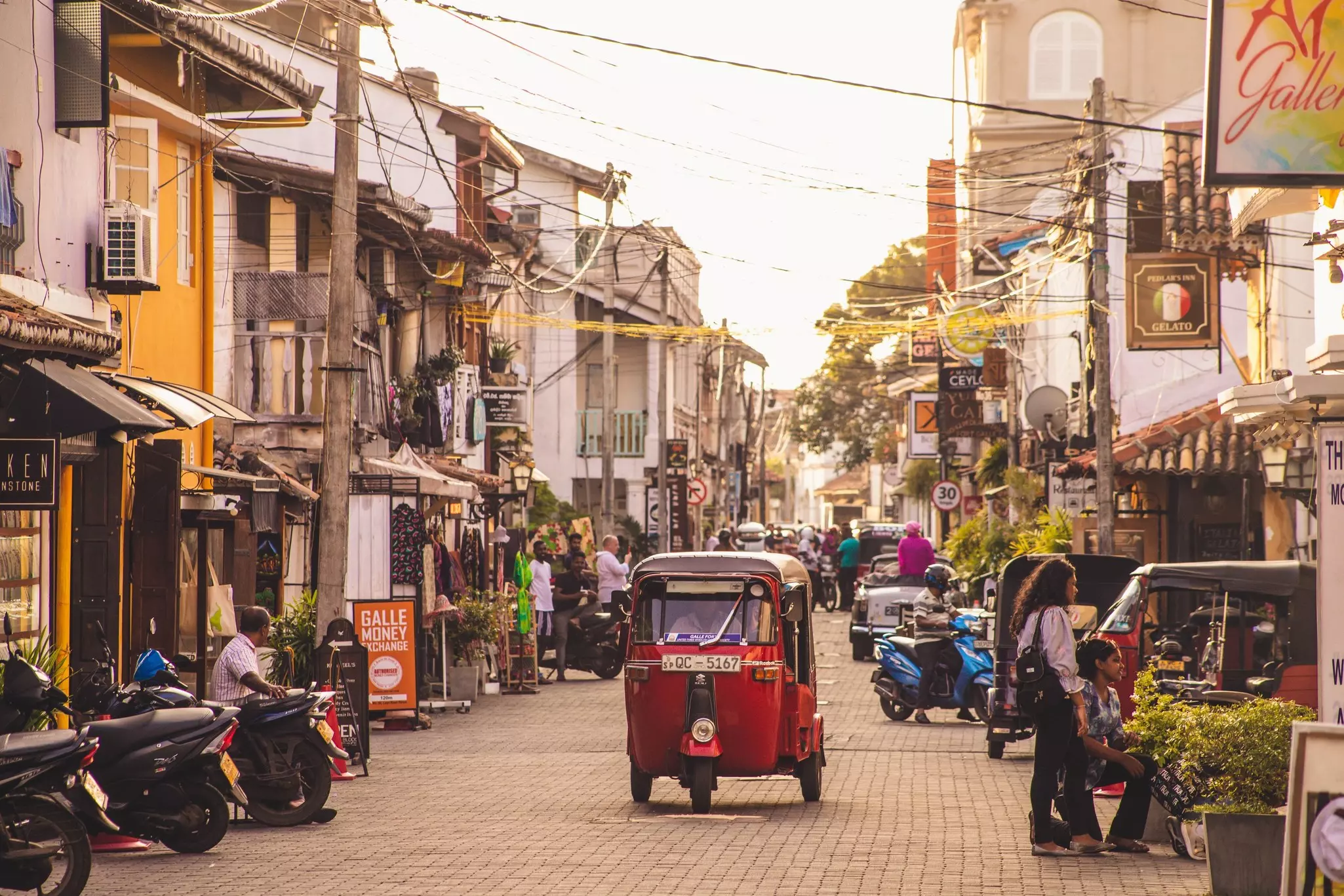A red tuk-tuk travels down a narrow street lined with shops in a town in the early evening in Galle Fort, Sri Lanka