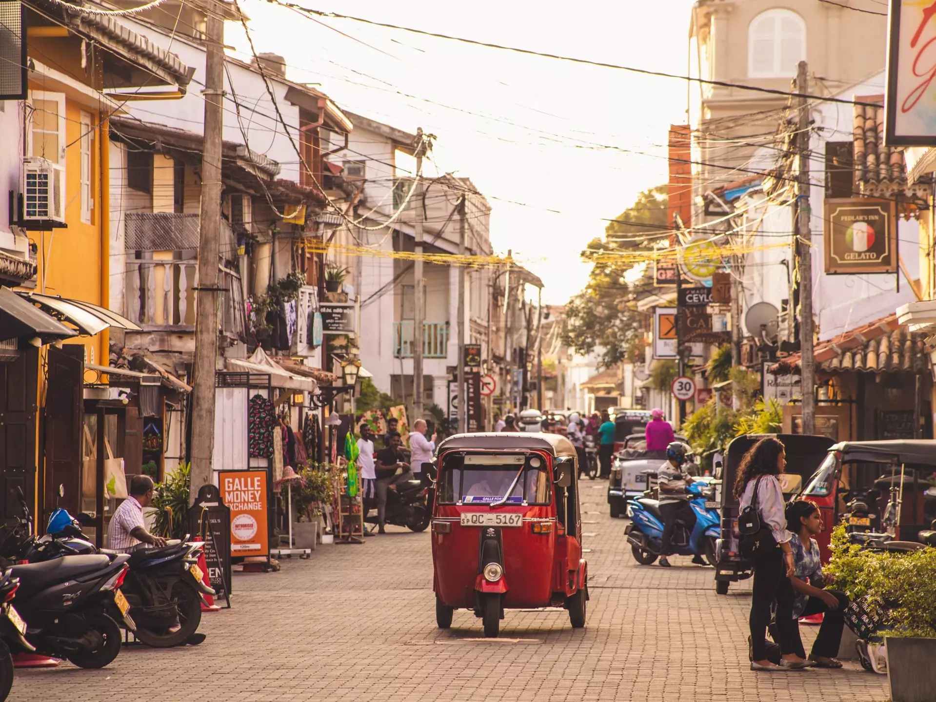 A red tuk-tuk travels down a narrow street lined with shops in a town in the early evening.