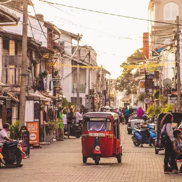 A red tuk-tuk travels down a narrow street lined with shops in a town in the early evening.