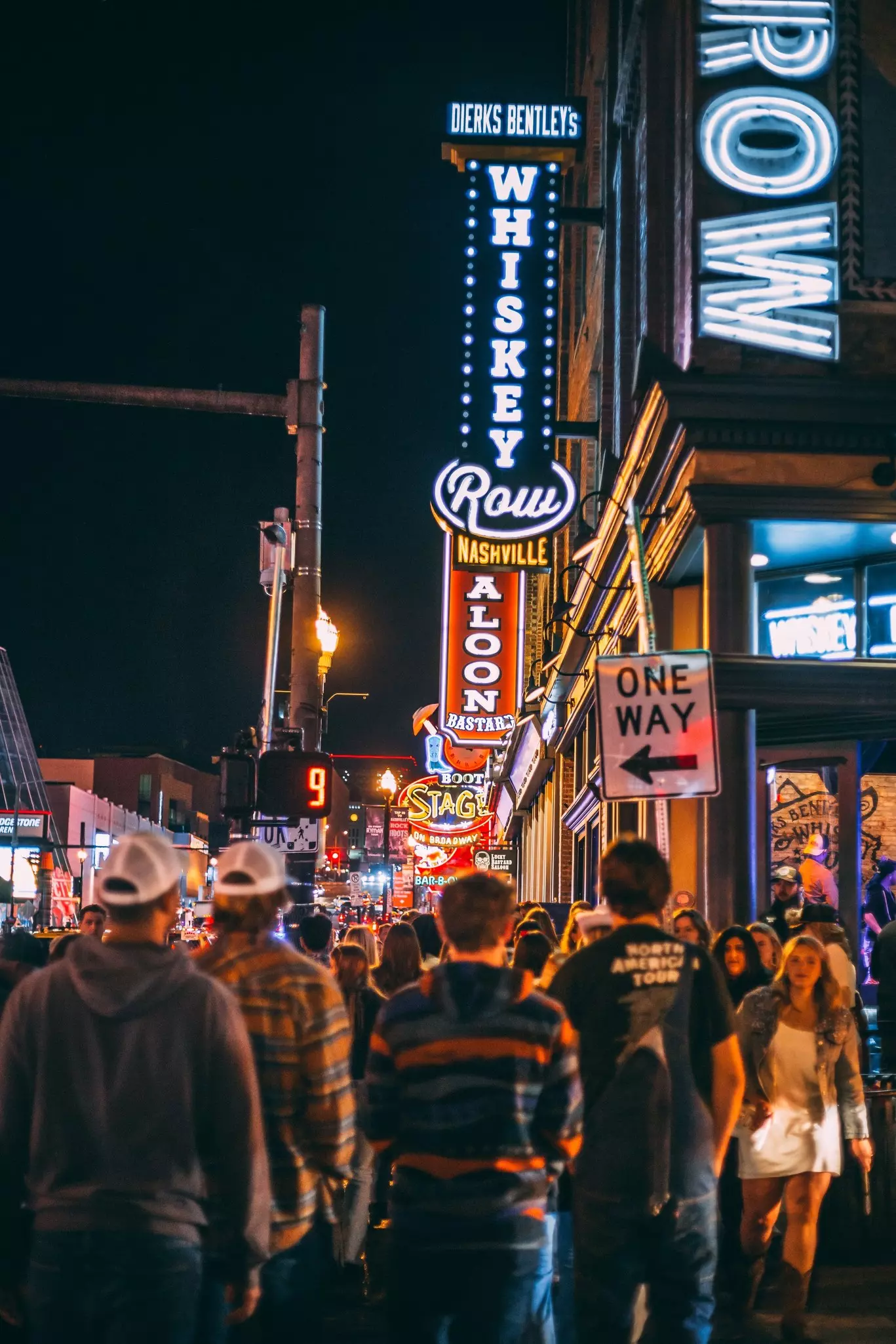 People walking on a crowded city street at night with neon signs reading "Whiskey Row" and "Nashville."