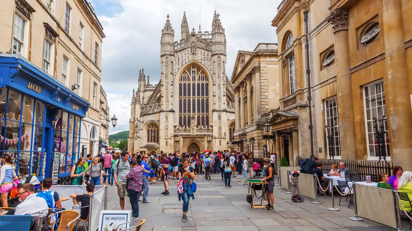 People walk in the street in front of the Bath Abbey, a towering church with a huge arched pane of stained glass facing the plaza.