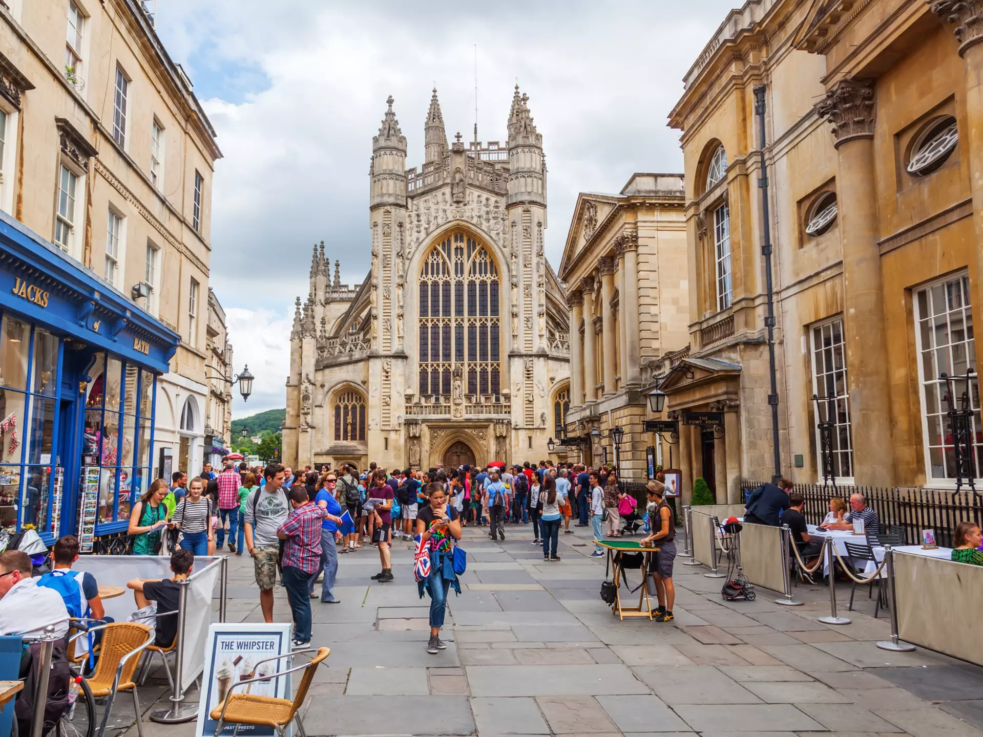 People walk in the street in front of the Bath Abbey, a towering church with a huge arched pane of stained glass facing the plaza.