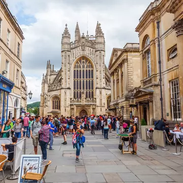 People walk in the street in front of the Bath Abbey, a towering church with a huge arched pane of stained glass facing the plaza.
