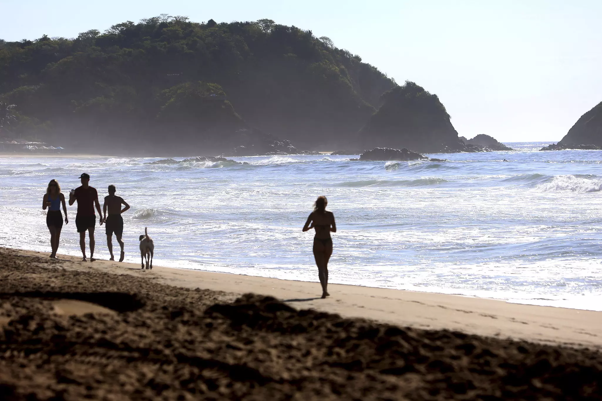 Four people and a dog in silhouette walking along a beach with a hilly headland in the distance.