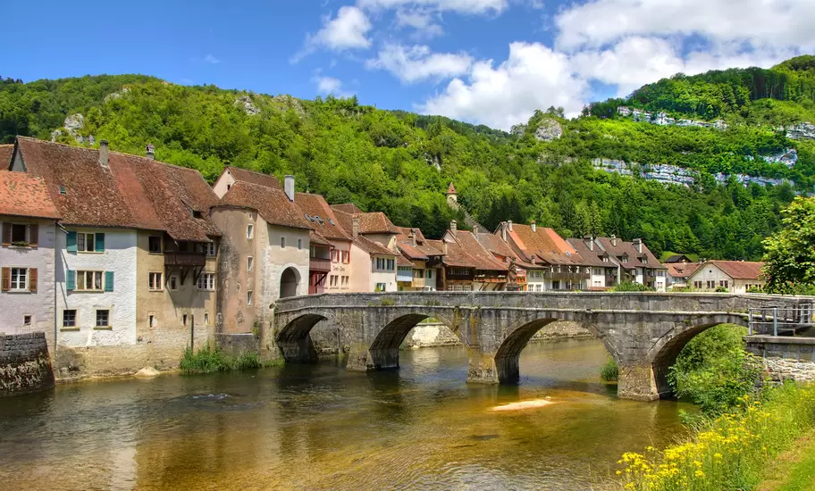 Arched stone bridge over the Doubs river at Saint-Ursanne, Switzerland, with old houses and wooded Jura slopes