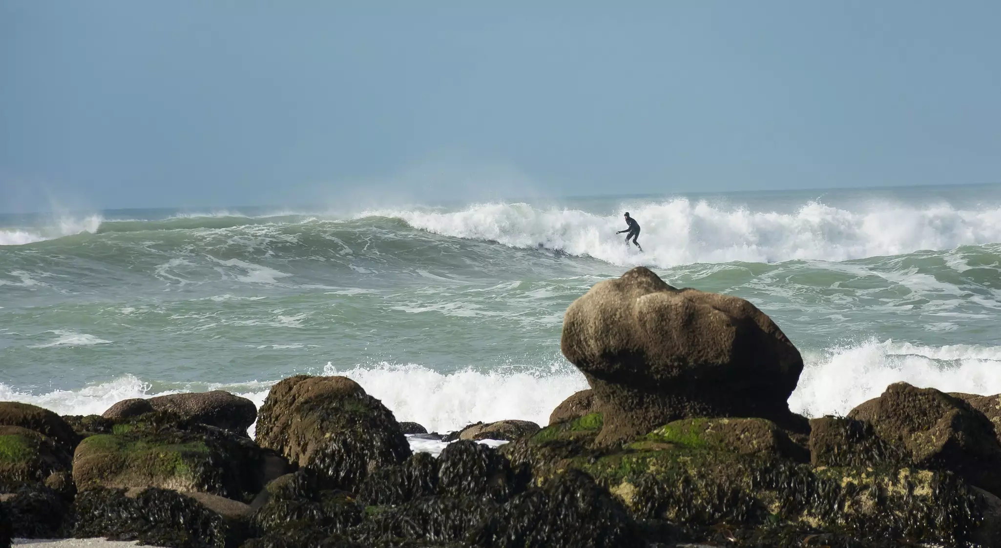A single surfer on a wave; rocks are in the foreground.
