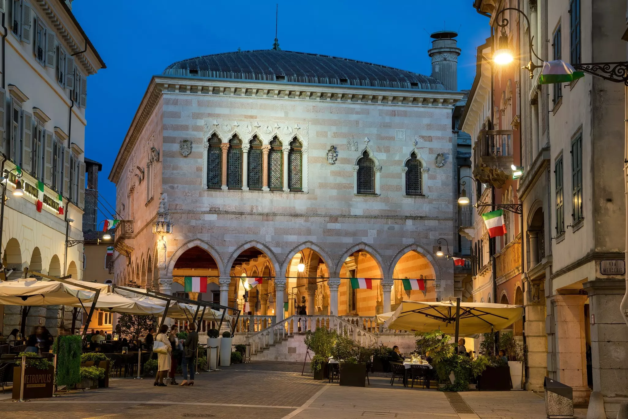 Outdoor tables covered by umbrellas with people standing nearby is pictured at dusk. A historic structure with pointed arches and windows is prominent just behind the plaza.