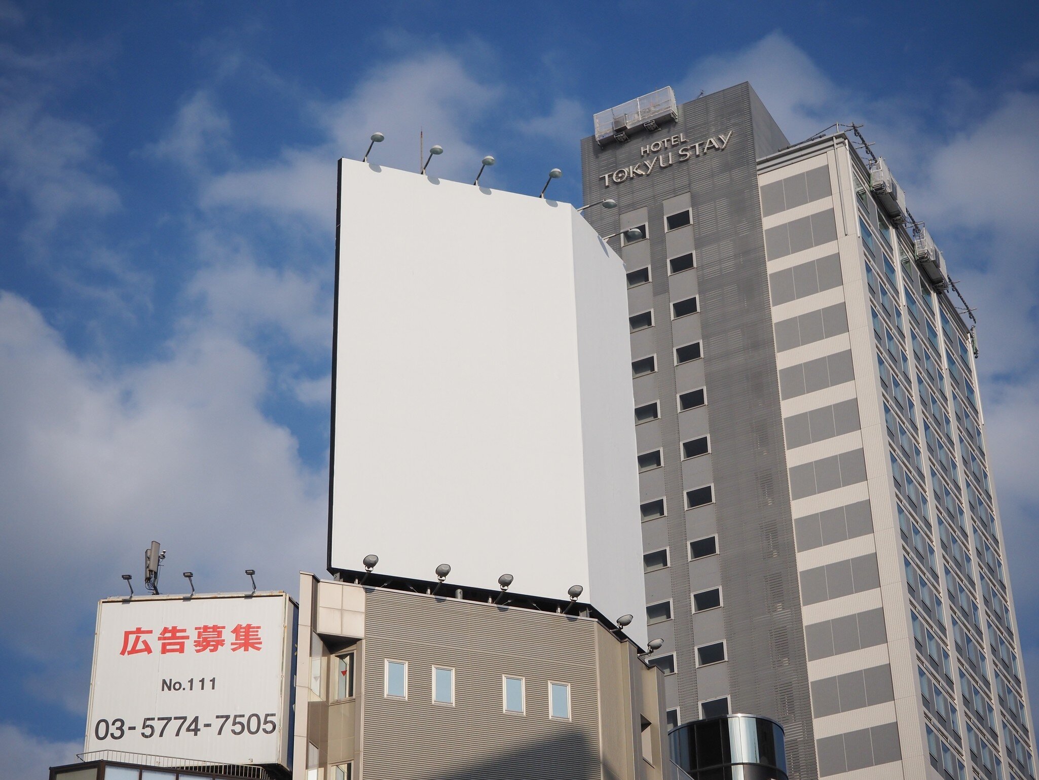 Tall, gray building with the words "Hotel Tokyu Stay" on it, rising into a blue sky