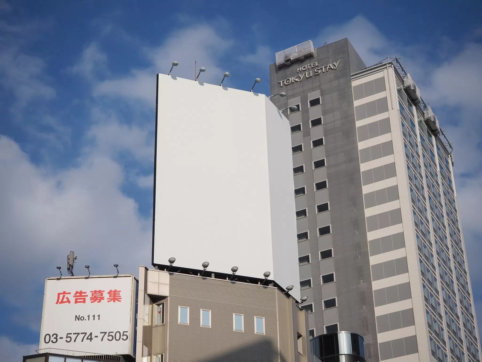 Tall, gray building with the words "Hotel Tokyu Stay" on it, rising into a blue sky