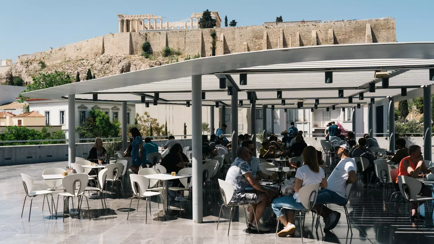 People at a semi-shaded outdoor cafe with ruins on a hillside in the distance on a sunny day.