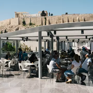 People at a semi-shaded outdoor cafe with ruins on a hillside in the distance on a sunny day.