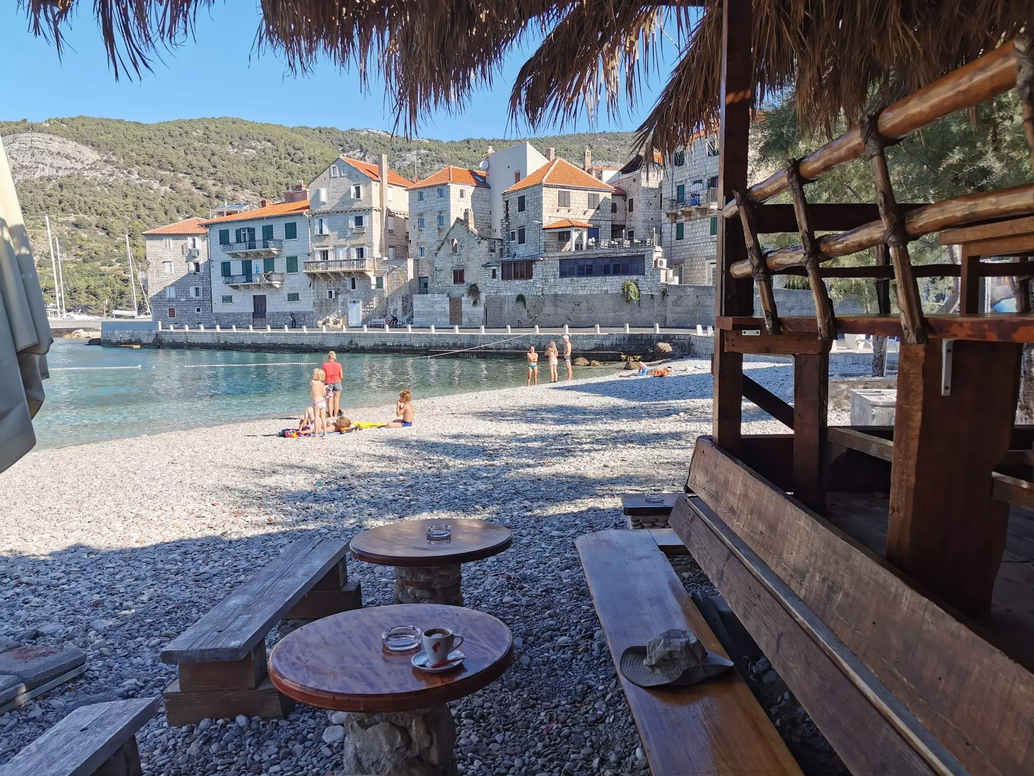 A stone dock lined with stone buildings along sparkling blue waters with children on the shore. In the forefront, a wooden bench with a sun hat on it, with a coffee on a small wooden table