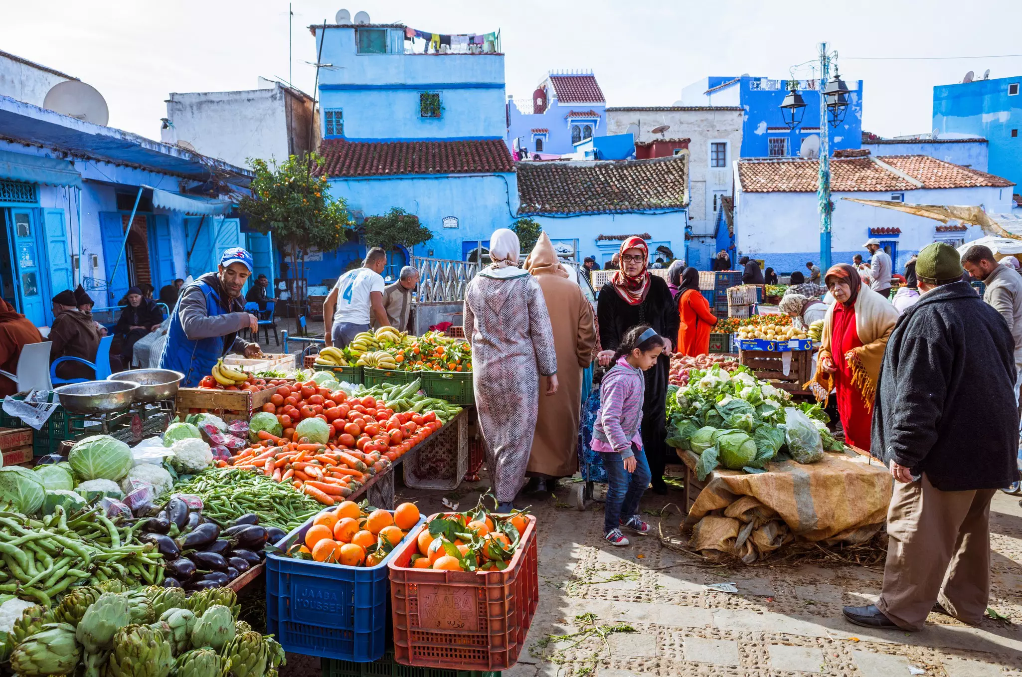 People shop for fruit and vegetables at a market square in Morocco; the buildings on the outer edge are washed in blue.