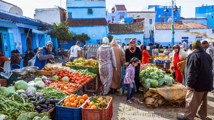 People shop for fruit and vegetables in a market square with blue buildings in the background. 