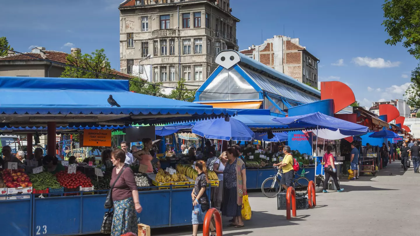 Shoppers stroll by a series of stalls packed with fruits and vegetables under a bright blue canopy.