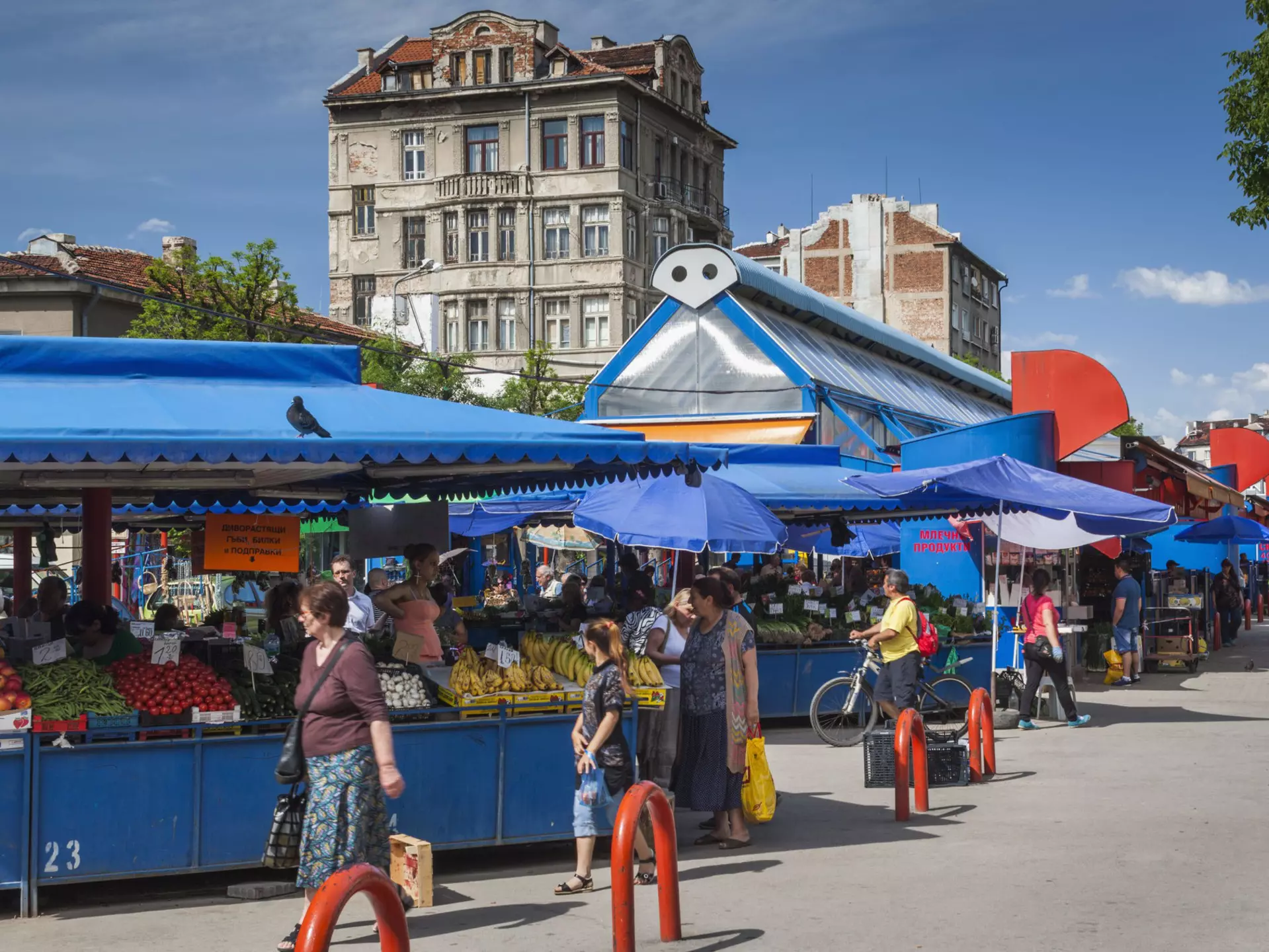 Shoppers stroll by a series of stalls packed with fruits and vegetables under a bright blue canopy.
