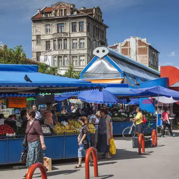 Shoppers stroll by a series of stalls packed with fruits and vegetables under a bright blue canopy.