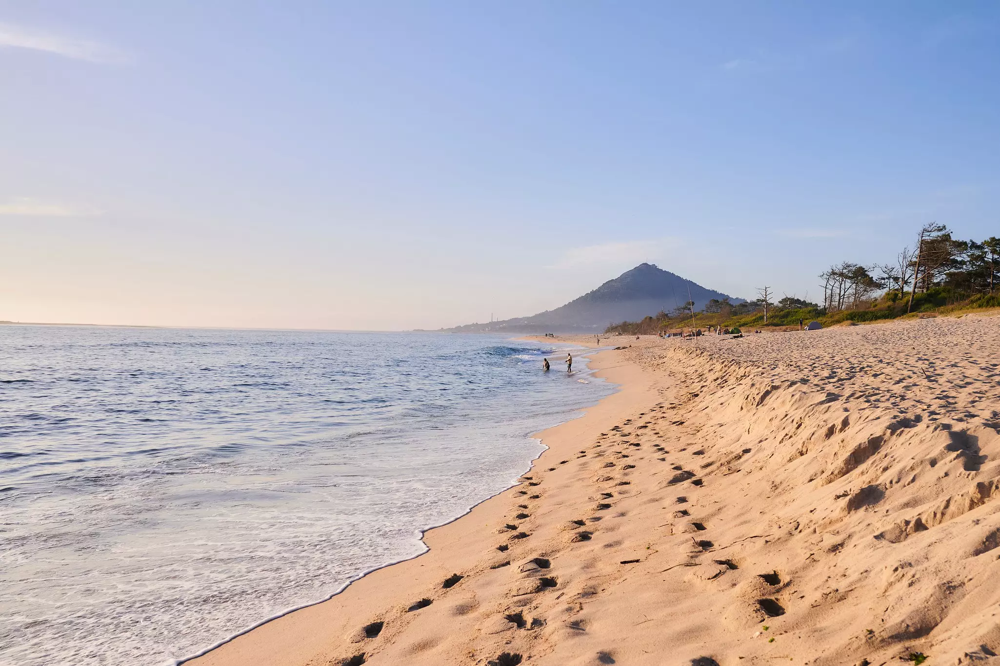 Two people at the edge of the surf at an early morning on a sandy beach. A pointed hill dominates the skyline in the distance.
