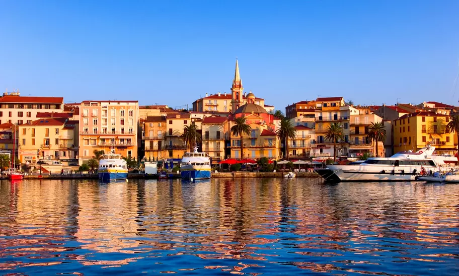 Calvi harbour in Corsica with pastel waterfront buildings, a church spire and dome, ferries and yachts reflected in the water