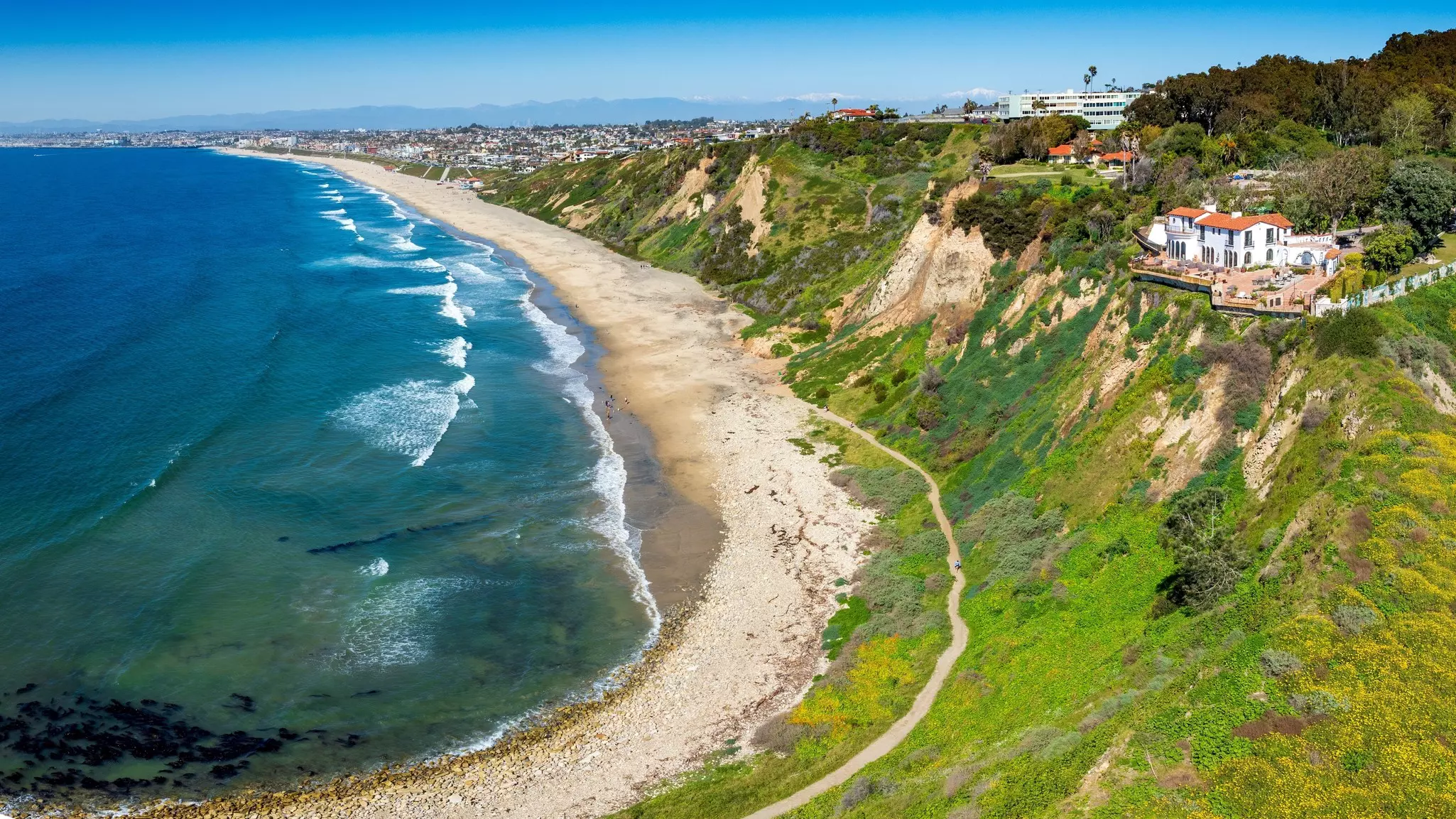 Aerial view of curved coastline beach with ocean to the left, homes on a hillside to the right, and a city view in the distance.