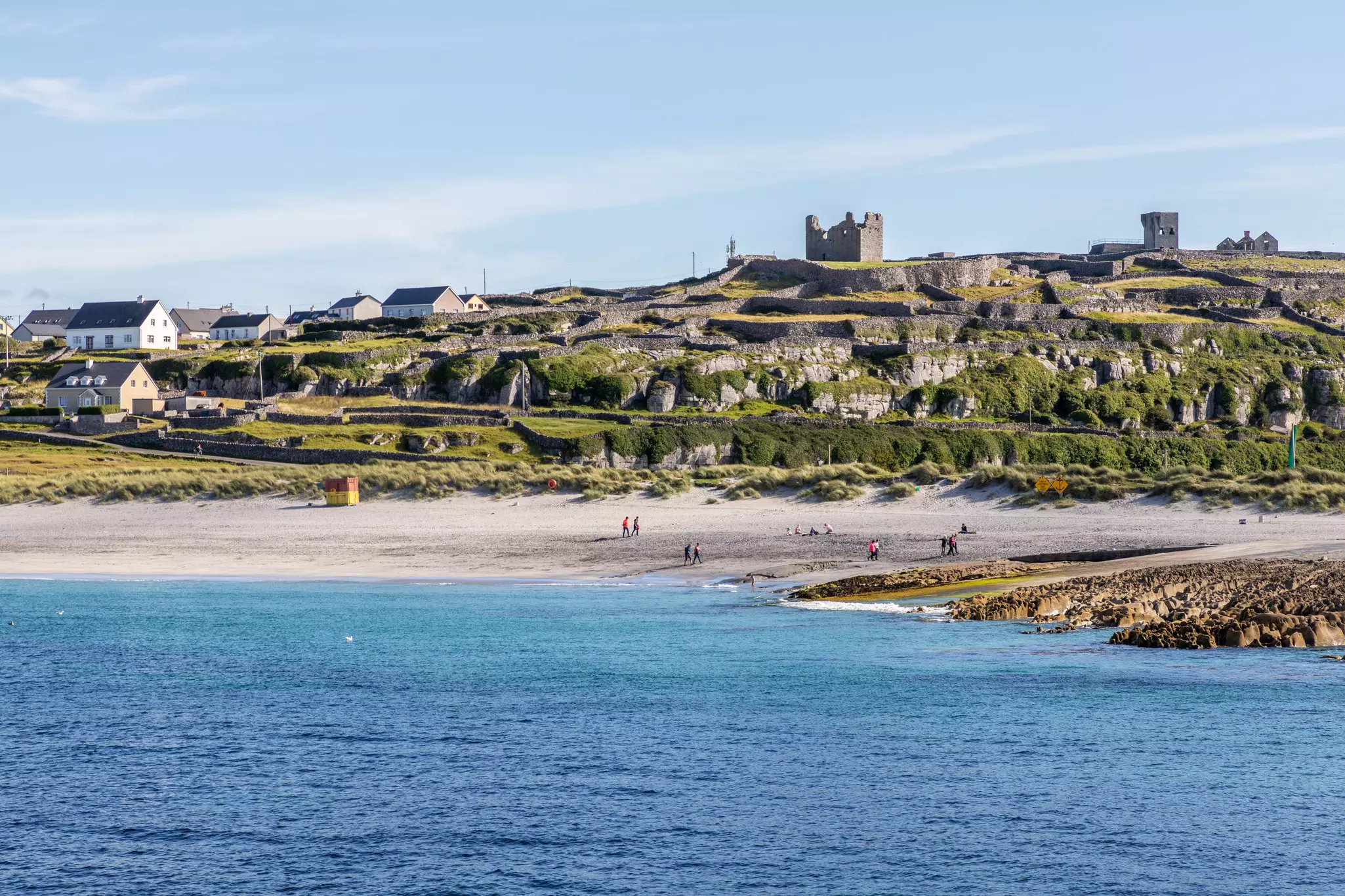 People walk along a beach on a sunny day. The beach is backed by a row of small houses and the ruins of a stone fort.
