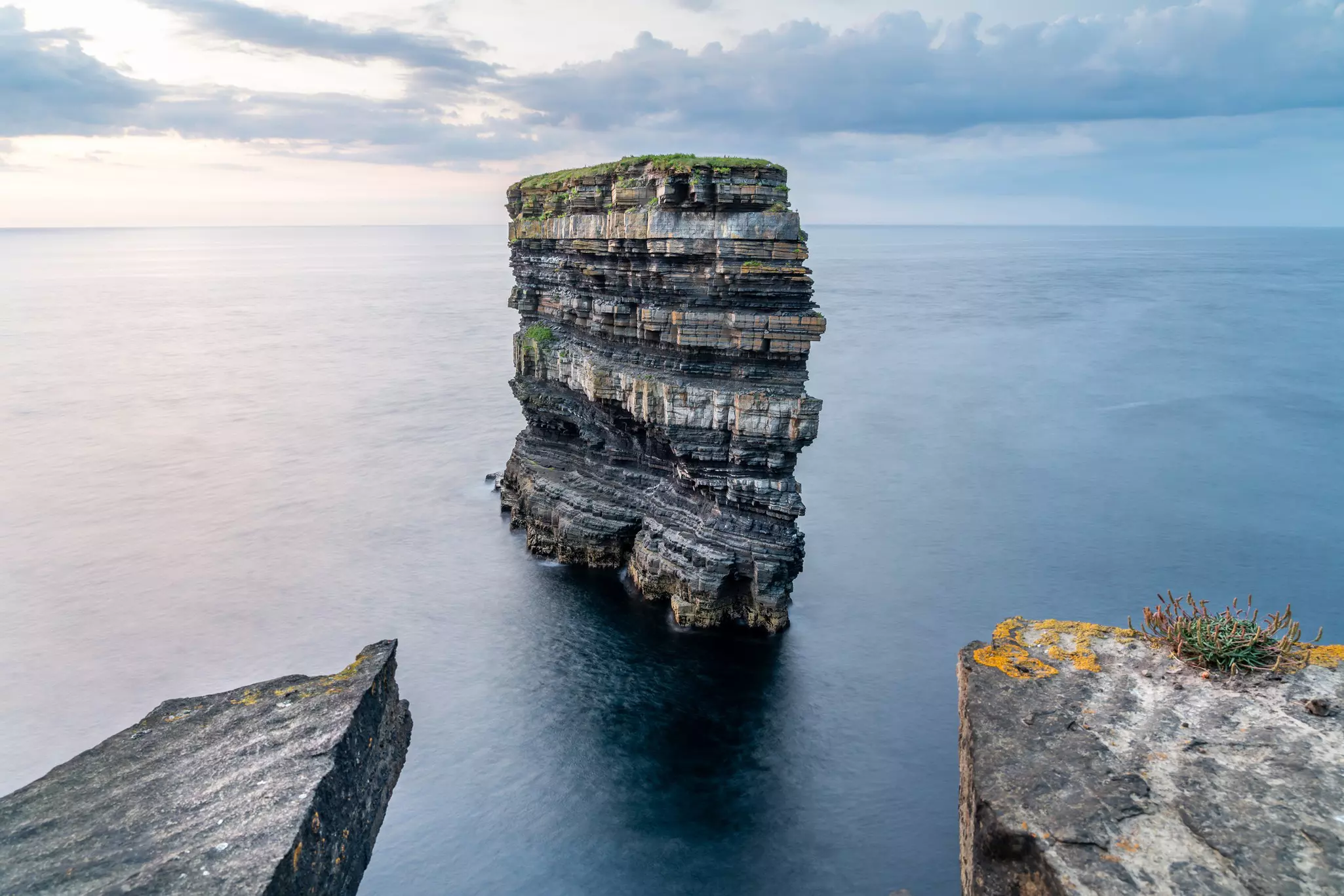 Waves crash at the base of the Dun Briste. There's bright green grass at the top of the natural formation on a dark cloudy day.