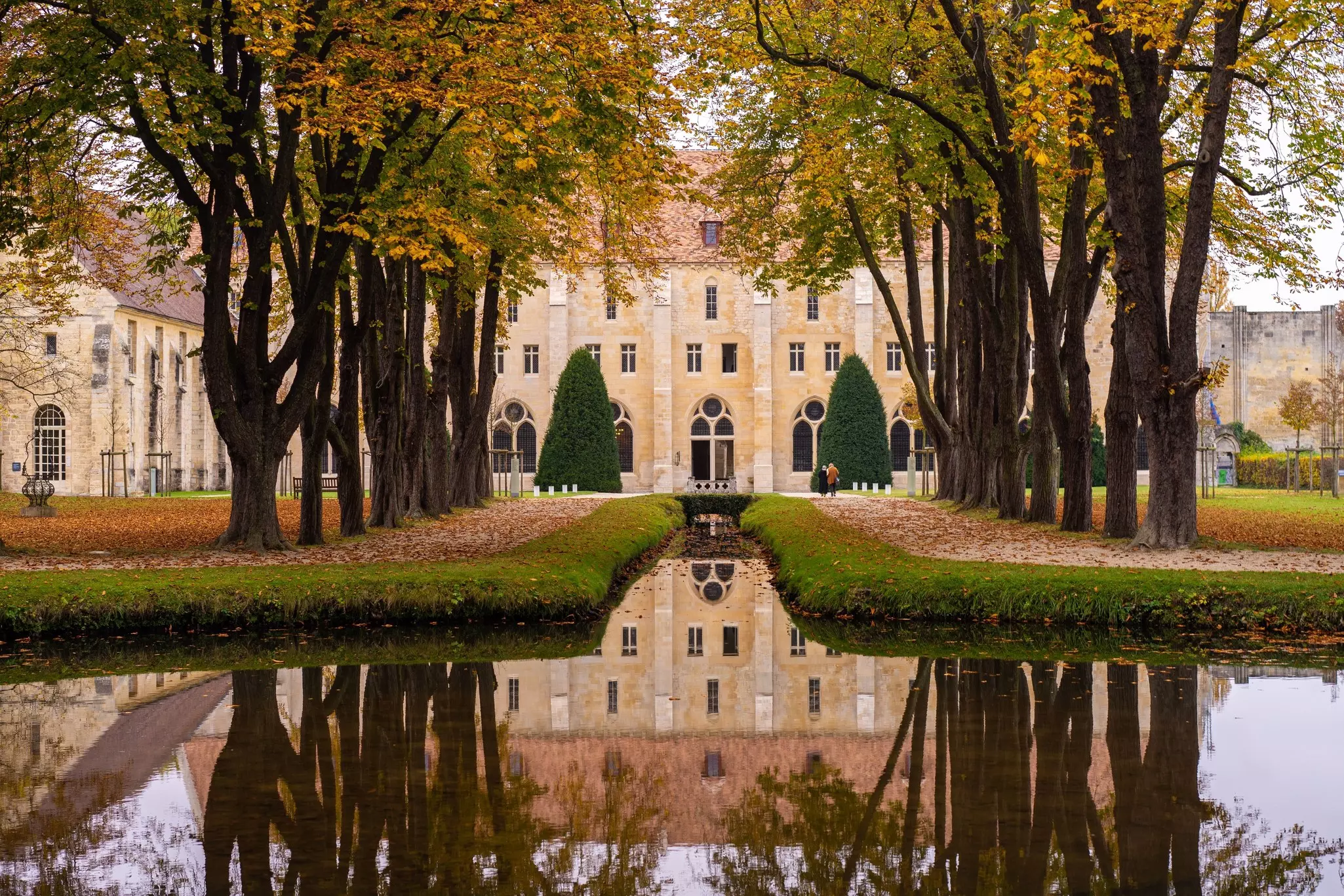 A grand abbey building in landscaped gardens with tree-lined paths and a central waterway.