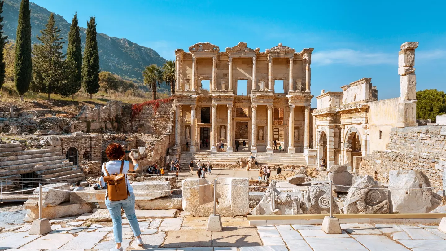 A visitor wearing jeans and a leather backpack takes photos of ancient ruins in Türkiye.