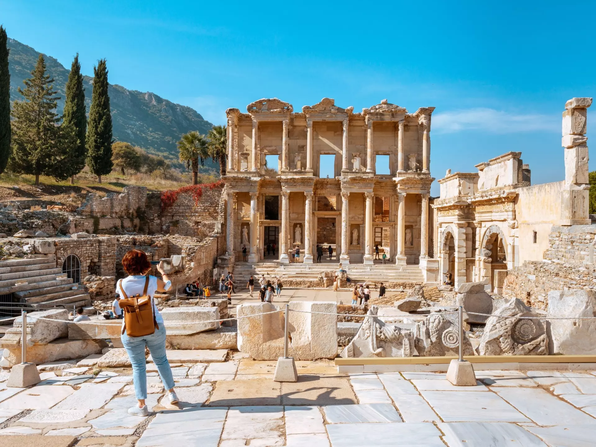 A visitor wearing jeans and a leather backpack takes photos of ancient ruins in Türkiye.