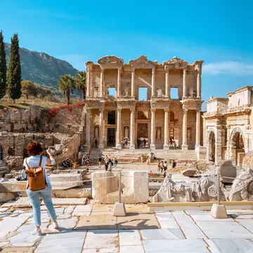 A visitor wearing jeans and a leather backpack takes photos of ancient ruins in Türkiye.