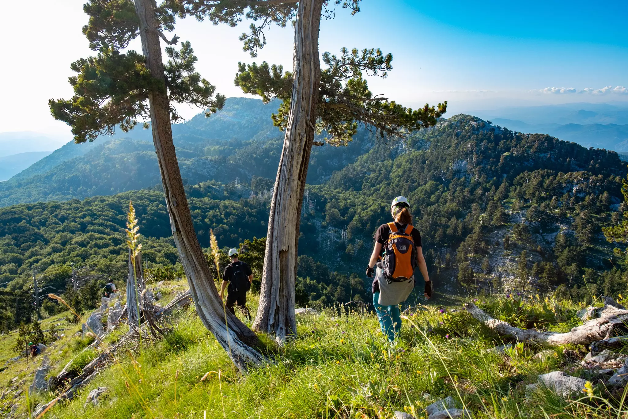Two hikers follow a trail through a mountainous region on a sunny day.