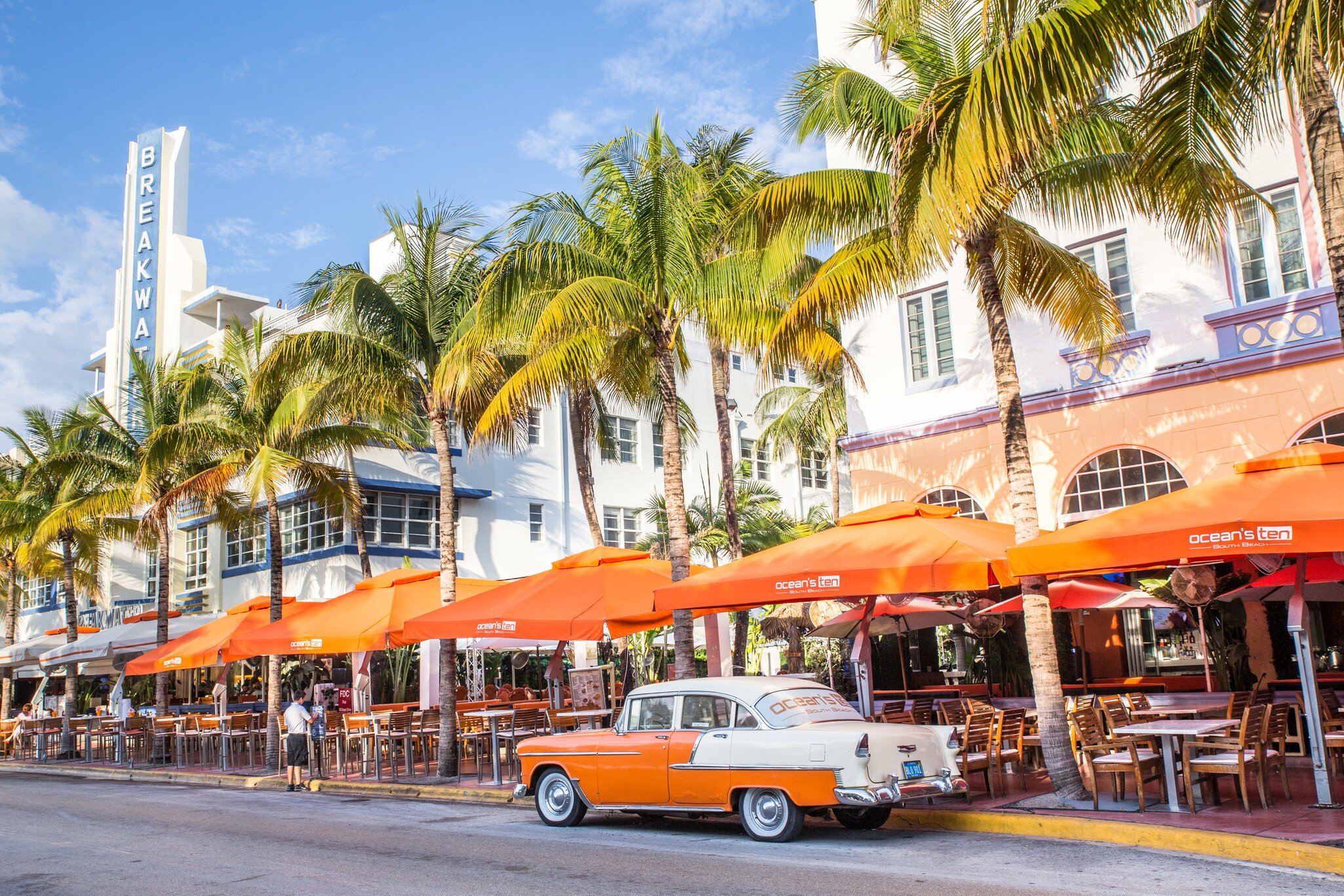 Orange and white vintage car on Ocean Drive beside palm trees and a white Art Deco hotel with a Breakwater sign, Miami.