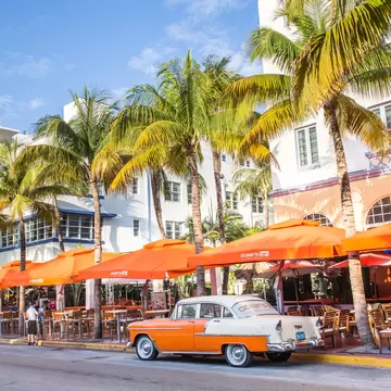 Orange and white vintage car on Ocean Drive beside palm trees and a white Art Deco hotel with a Breakwater sign, Miami.