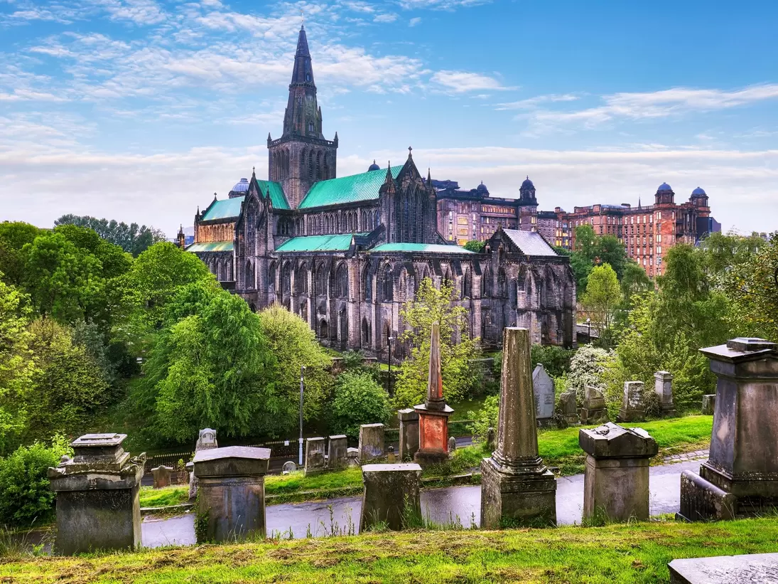 A large Gothic cathedral viewed from a hilltop covered in graves and monuments: Glasgow Cathedral and Glasgow Necropolis on a rainy day.