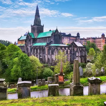 A large Gothic cathedral viewed from a hilltop covered in graves and monuments: Glasgow Cathedral and Glasgow Necropolis on a rainy day.