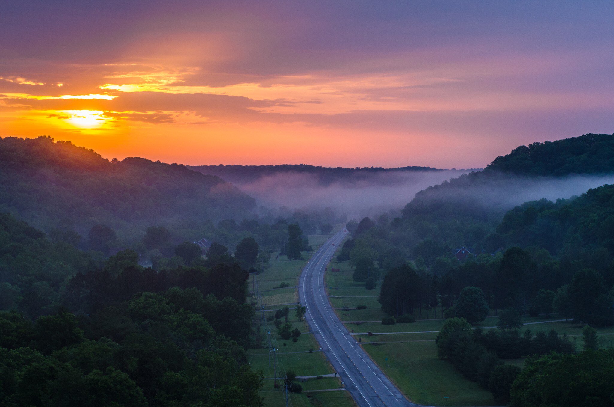 A two-lane highway curving through misty forested hills under an orange sky.