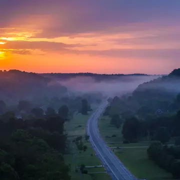 A two-lane highway curving through misty forested hills under an orange sky.