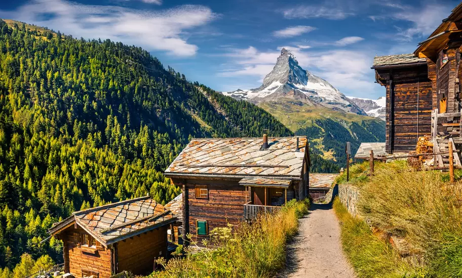 Weathered wooden chalets with stone-slab roofs line a mountain path below the snowy Matterhorn in Valais, Switzerland