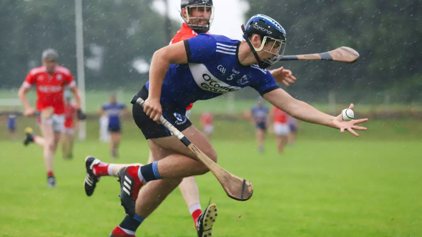 A man in a blue jersey and shorts is holding a hurl and sliotar as a man in a red jersey and shorts attempts to tackle him.