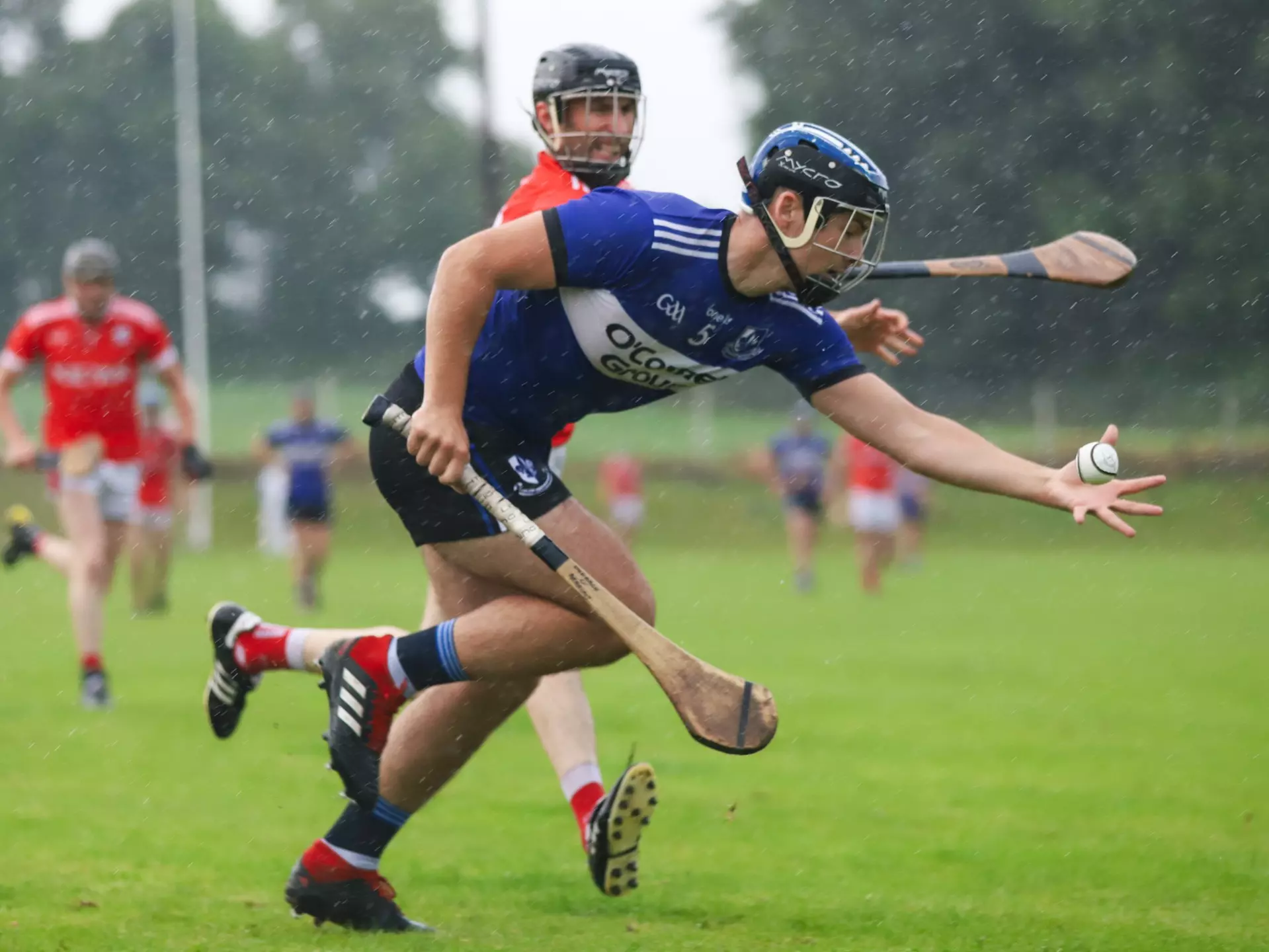 A man in a blue jersey and shorts is holding a hurl and sliotar as a man in a red jersey and shorts attempts to tackle him.