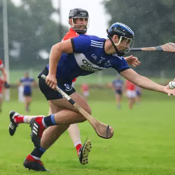 A man in a blue jersey and shorts is holding a hurl and sliotar as a man in a red jersey and shorts attempts to tackle him.
