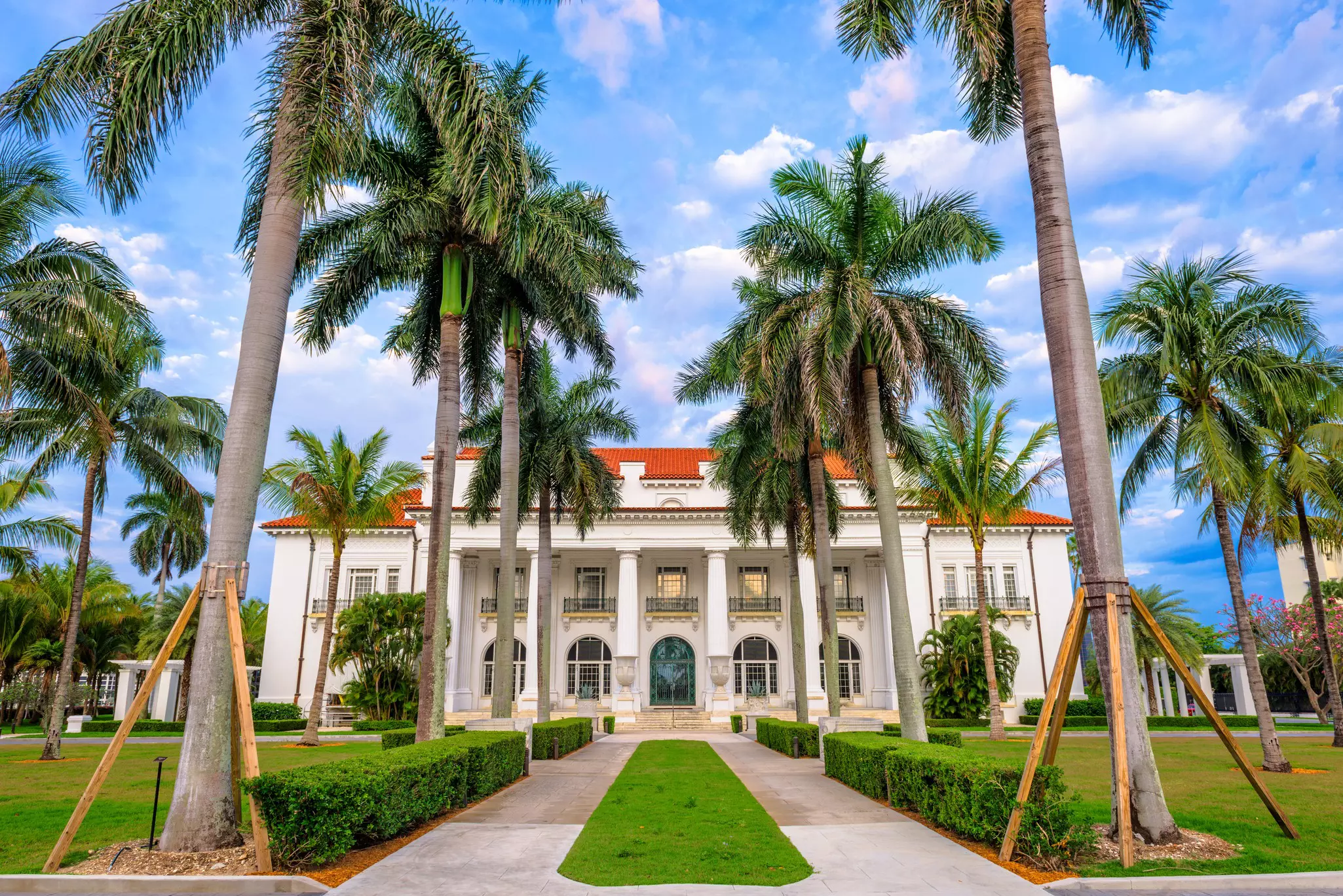 A driveway lined with palm trees leads to a mansion, with ornate columns and a red-tiled roof.