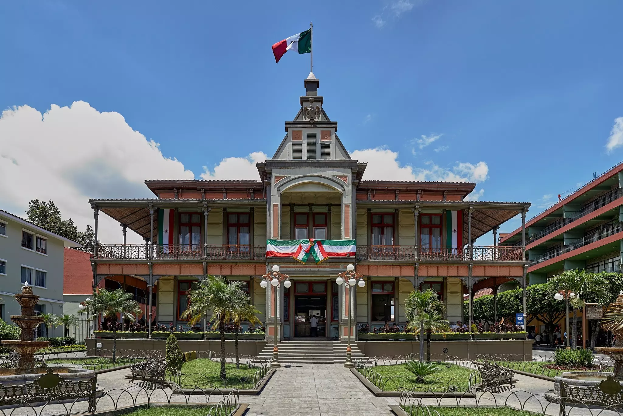 A historic building with a covered porch and balcony seen from a formal garden square on a sunny day.