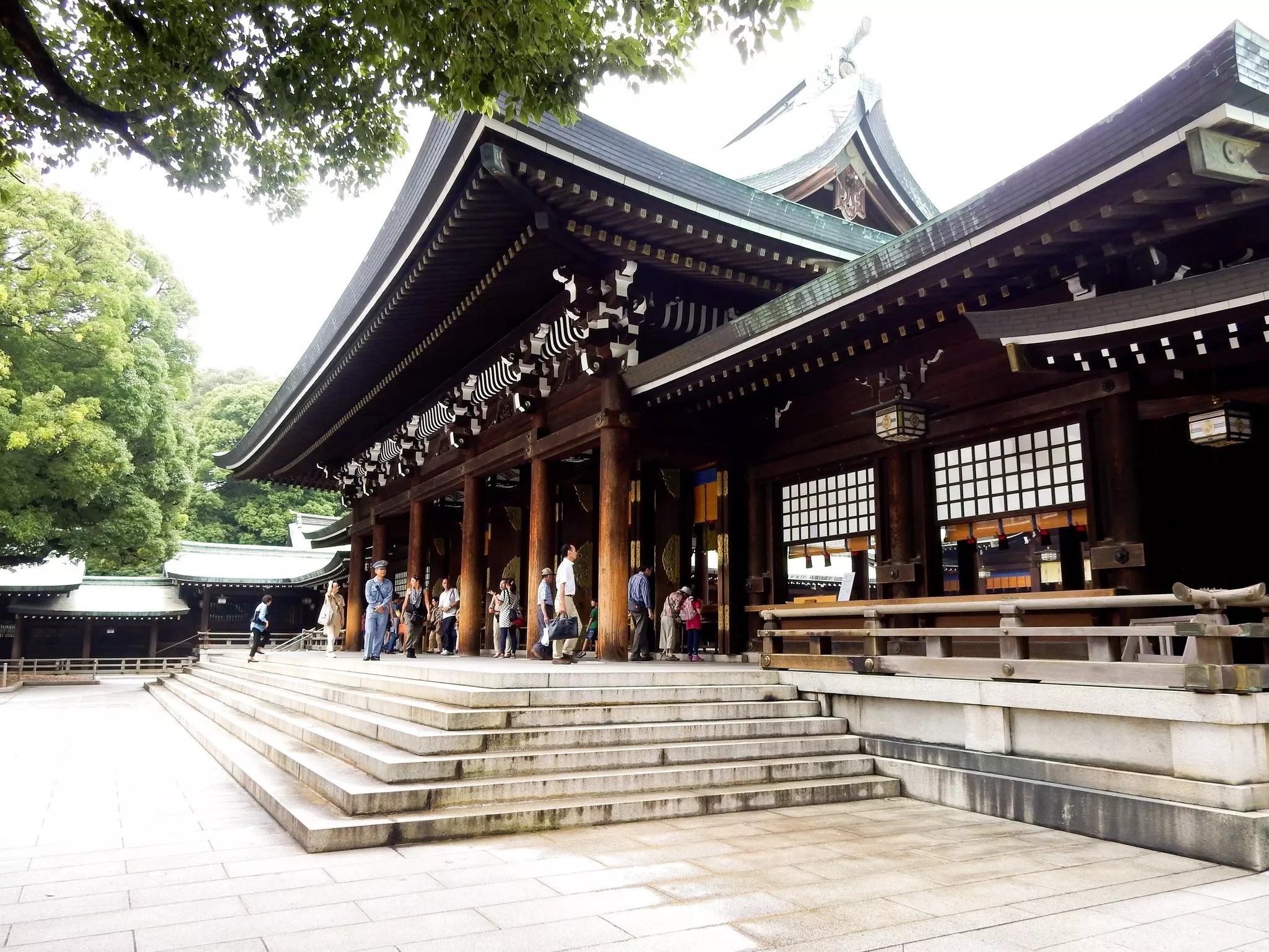 People enter the pavilion of a temple. The brown-wood structure has overhanging roofs with flared eaves.