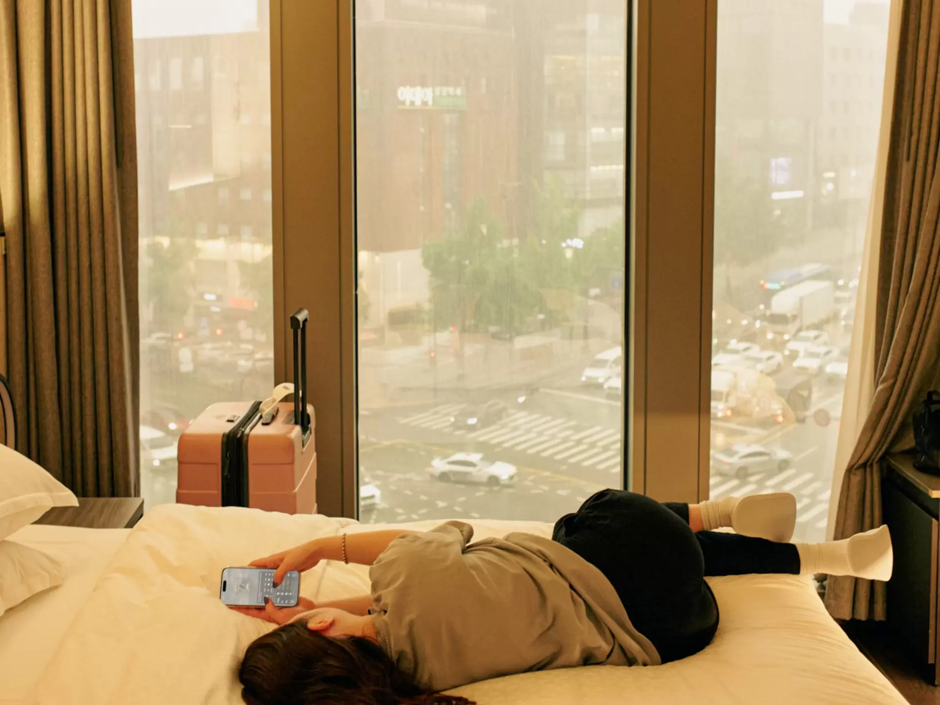A woman lies on her hotel room bed checking the weather on her phone, with a city skyline behind her. Her suitcase stands upright on the other side of the bed by the window