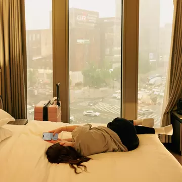 A woman lies on her hotel room bed checking the weather on her phone, with a city skyline behind her. Her suitcase stands upright on the other side of the bed by the window