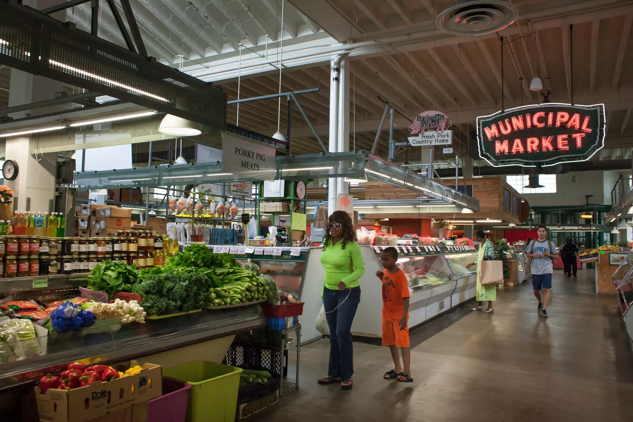 People shop at food stalls at an indoor market, with a neon sign overhead.