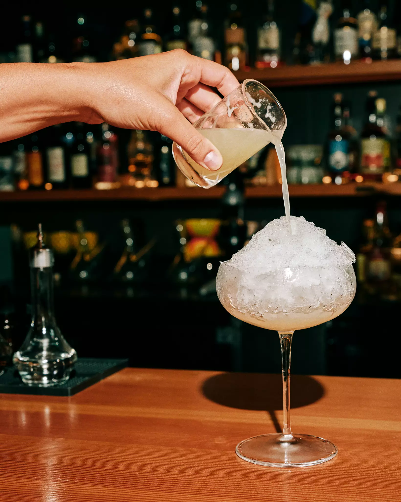 A hand pours a drink from a small beaker into a ice-filled coupe glass.