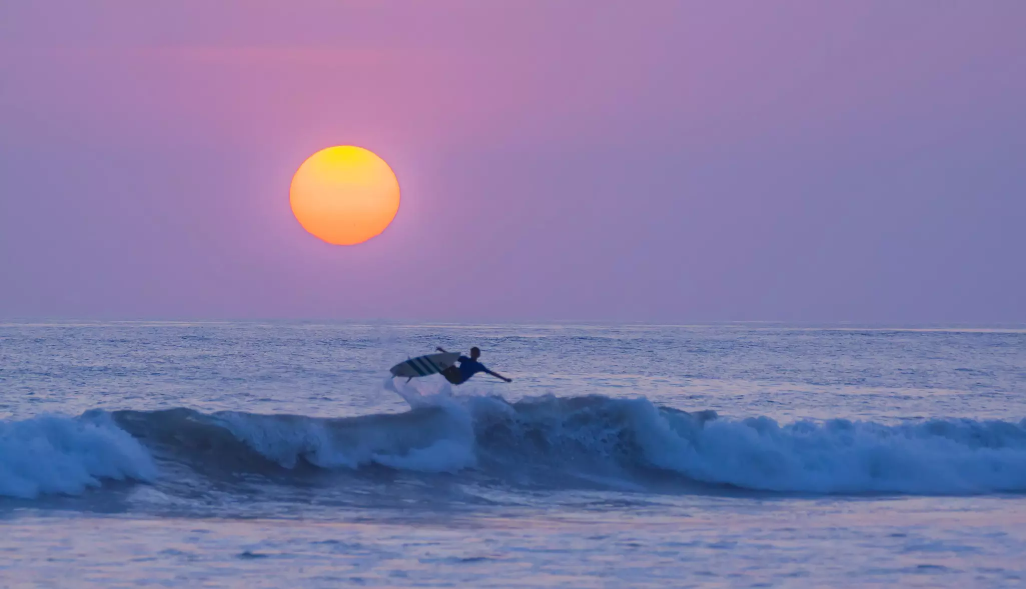 A person surfs over a wave with the sunrise or sunset in the background